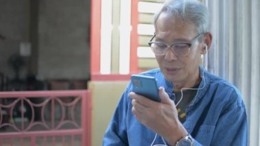Asyalı mutlu son sınıf gri saçlı baba evinin önünde otururken kulaklıkla video bağlantısı kuruyor. Modern teknoloji. Yaşlı olgun adam sağlıklı yaşam tarzı.