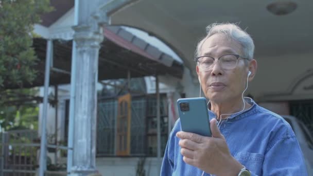 スマートフォンで会話をしているイヤホンを装着したメガネ姿のシニア白髪の男性が 自宅の前に立っている間 モバイルアプリ経由でビデオ通話で友人に肖像画 社会的距離 — ストック動画