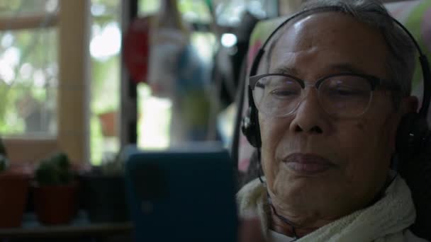 戴着耳机的戴眼镜的亚洲退休老人喜欢在房间里听音乐或看手机上的社交视频 放宽长者健康的生活方式 — 图库视频影像