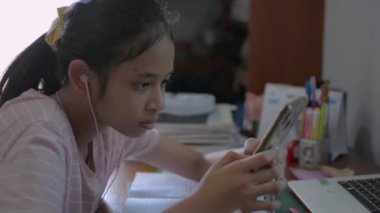 Asyalı ortaokul öğrencisi kulaklık takıyor. Evdeki internet dersinde akıllı telefonlu uzaktan öğretmene mesaj yolluyor. Yeni normal yaşamla çalış. 