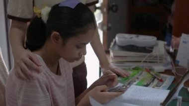 Asyalı genç kız internet sitesinde bilgi aramak için cep telefonunu kullanırken ablası ev işi yaparken içeri girip tavsiye veriyor. Çevrimiçi talimatlar. Evde eğitim.