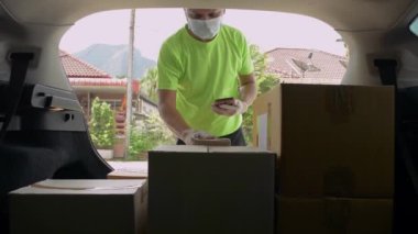 Yeşil tişörtlü Asyalı teslimatçı paket siparişleri listesini mobil cihazla kontrol ediyor ve evdeki bayan müşteriye gönderilen araçtaki bir kutuyla ilgileniyor. Bir paket almak için akıllı telefonu imzalıyorum. Bir arabanın içinden görüntüle.