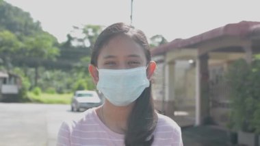 Oldukça uzun saçlı Asyalı genç kız evinin önünde maske takıyor. COVID-19 salgını sırasında koruyucu maskeli Taylandlı bir kızın portresi. Kameraya bakıyorum..