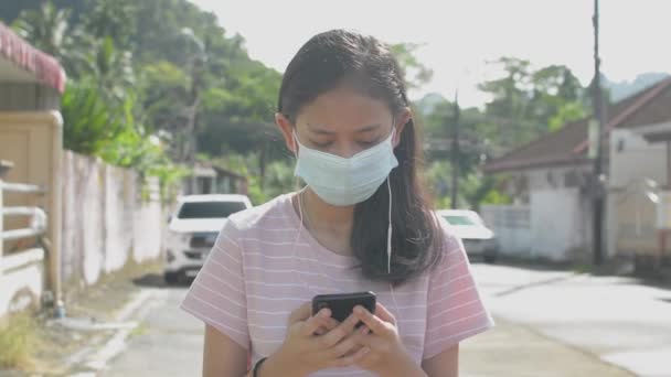 照片中的泰国长发漂亮女孩站在住宅区的街道上 戴着防护面具 从智能手机和耳机在线观看社交媒体 看着相机 — 图库视频影像