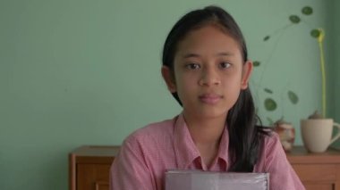 Asyalı mutlu uzun saçlı kız, kameraya bakarak gülümsüyor. Taylandlı genç kız öğrenci evde dururken elinde bir ders kitabı tutuyor. COVID-19 salgını sırasında evde kal.