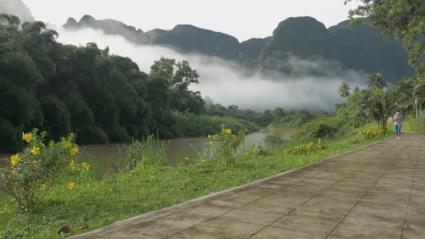 霧深い山のある川の美しい景色を眺めながら アジアの女性は午前中に歩道をジョギングするスポーツウェアでフェイスマスクを着用しています 田舎の風景 健康的なライフスタイル 公害のない環境 — ストック動画