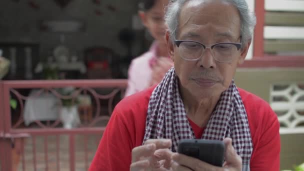 亚洲可爱的孙女走近她 兴奋地对她的祖父耳语着 而他却在家里用手机 两代人在一个家庭的结合 快乐与团结 — 图库视频影像