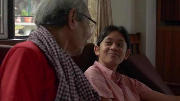 可爱的亚洲少女孙女坐在客厅里和她的长发祖父聊天时 开心地笑着 两代人在一个家庭的结合 老年人与儿童的关系 — 图库视频影像