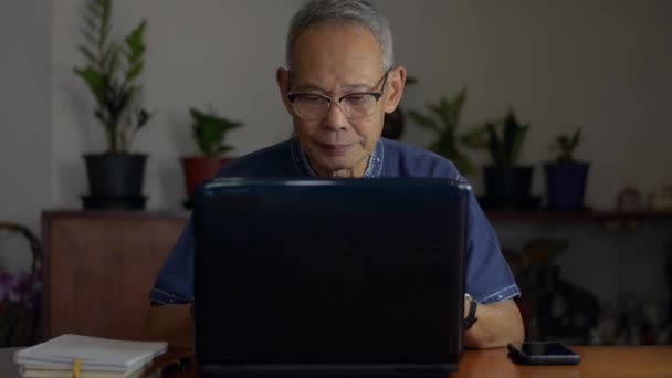 アジア系のシニア定年退職者は 机の上のノートパソコンで眼鏡をかけ 自宅のノートパソコンに書き込みます 家から仕事だ テクノロジーと高齢者のライフスタイルの概念 — ストック動画