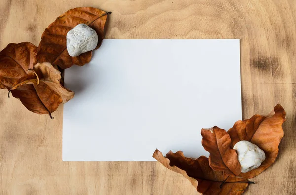 Mockup. A white sheet of paper on a wooden surface, surrounded by autumn leaves and white stones. Selective focus.