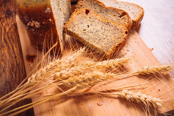 Scheibe frisches Brot mit Mohn und Weizenähren auf Holzgrund schließen — Stockfoto