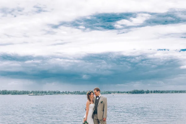 Молодой жених и невеста стоят, обнимаясь на фоне реки — стоковое фото