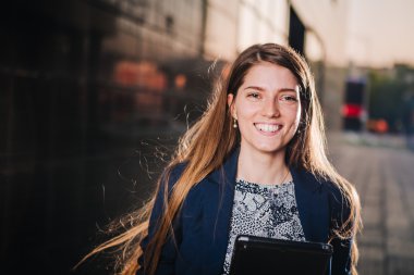 Successful beautiful young business woman is smiling on the background of buildings and holding a tablet computer.