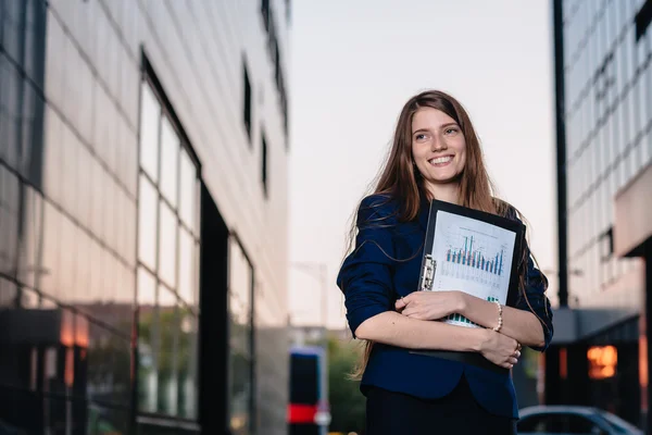 Successful smiling businessman, standing against the backdrop of buildings holding  folder with sales charts. City business woman working. — Stockfoto