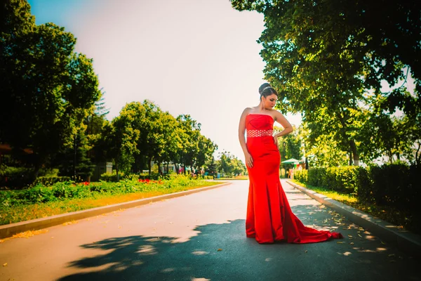 Beautiful girl in a long red evening dress standing on the background of trees and shrubs — Stock Photo, Image