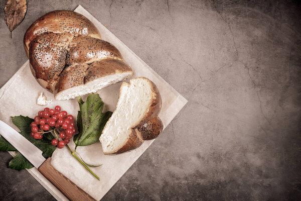 On the table is a sliced loaf of white bread with a crisp crust sprinkled with poppy seeds. Next to the knife and viburnum berries. Front view, dark background, copy space