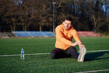 Spor sahasında oturan ve dışarıda egzersiz yaparken bacağını esneten bir adam. Fitness, ısınma ve aktif yaşam tarzı