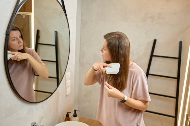 Evde banyo aynasının önünde duran bir kadın uzun saçlarını tarıyor. Günlük tımar kavramı