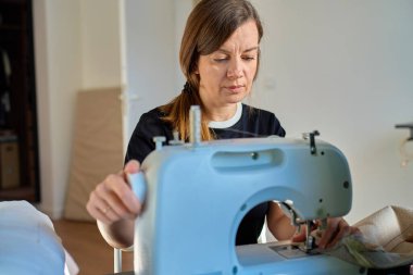 Evde oturan bir kadın dikiş makinesi kullanıyor ve kumaşa elleriyle rehberlik ediyor. Ev dikimi kavramı