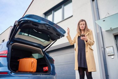 Açık bagajlı, turuncu çantalı, bej ceketli bir kadın, telefonu tutuyor ve elini kaldırıyor.