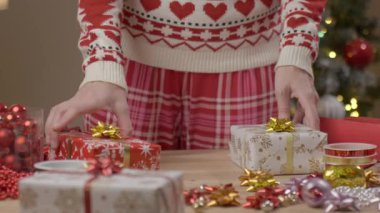 Noel süveteri giyen bir kadın hediye kutularını tahta masada kurdele kurdelesiyle düzenliyor. Tatil hazırlığı ve hediye sunumu kavramı