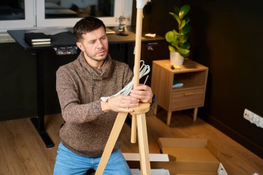 Evin içinde elektrik kablosu ve paketlenmemiş kutularla yer lambasını birleştiren bir adam. Mobilya montaj kavramı