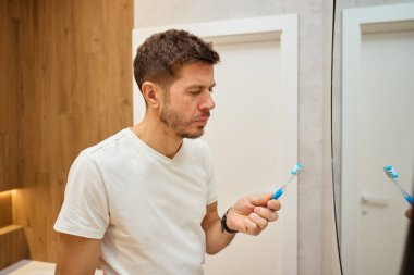 Adam banyoda dikilip aynaya bakarken diş fırçasına bakıyor. Ağız hijyeni ve diş bakımı kavramı