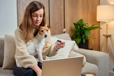 Kanepede oturan bir kadın evde kredi kartı tutarken dizüstü bilgisayar kullanıyor, köpek kucağında. Çevrimiçi alışveriş ve dijital ödeme kavramı