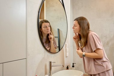 Lavaboda duran ve banyo aynasında yüzünü inceleyen bir kadın. Cilt rutini kavramı