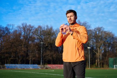 Spor sahasında dikilirken elinde su şişesi tutan turuncu ceketli bir adam. Susuzluk ve zindelik eğitimi kavramı