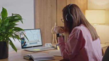 Woman wearing headphones, drinks coffee and begins writing in notebook while sitting at desk with laptop. Concept of online learning, remote work