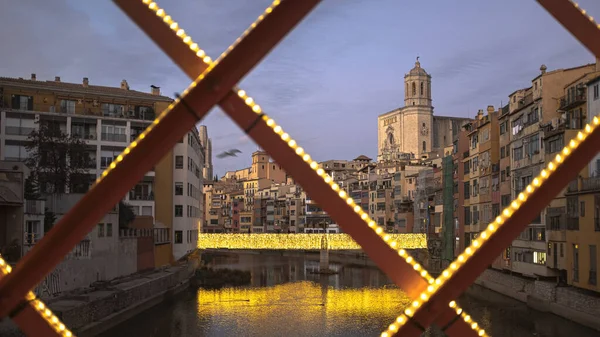 Girona 'nın kentsel manzarası alacakaranlıkta gökyüzü ünlü Gotik katedral simgesi ve nehir evleriyle birlikte Red Iron Köprüsü' nden gelen sessiz bir nehri yansıtıyor.