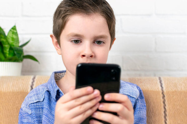 Эмоциональный мальчик с серьезным лицом, сосредоточенный на телефоне. Ребенок смотрит в смартфон, печатает и отправляет сообщение, играя в игру дома. Технологии, интернет-коммуникации, отдых для детей.