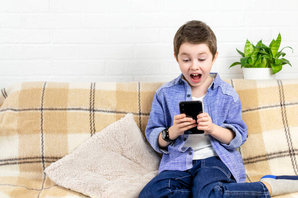 Эмоциональный мальчик пользуется телефоном. Ребенок удивил лицо и открыл рот, шокированный школьник смотрит в смартфон дома. Технологии, интернет-коммуникации, отдых для детей и людей.