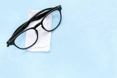 Siyah çerçeveli gözlük ve temiz bez. Mavi arka planda fotokopi alanı var. Kir ve tozdan gözlük silmek için beyaz yumuşak doku. Görüş düzeltme, optik bakım, göz hekimi, optometri.