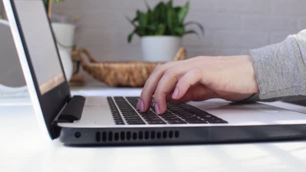 Kvinnliga händer av en affärskvinna eller student skriva på en bärbar tangentbord och göra anteckningar med en penna i en anteckningsbok vid en vit hemmakontor bord, online-utbildning, frilans, närbild, sidovy — Stockvideo