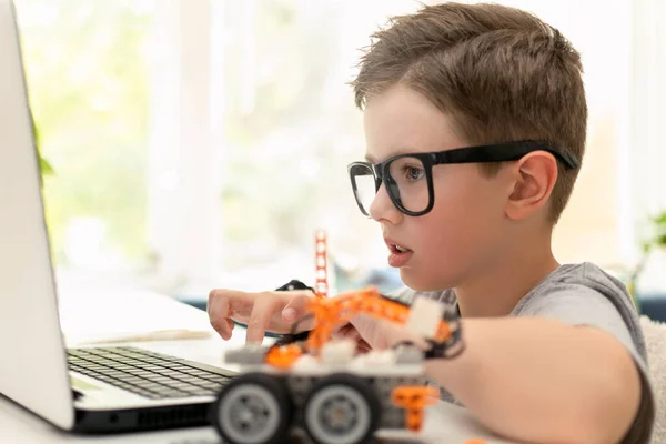 Un chico inteligente construye un coche robot usando un portátil cerca de la ventana en casa. El niño está aprendiendo programación y codificación. Robótica, ciencia, matemáticas, ingeniería, tecnología. Educación STEM para niños. — Foto de Stock