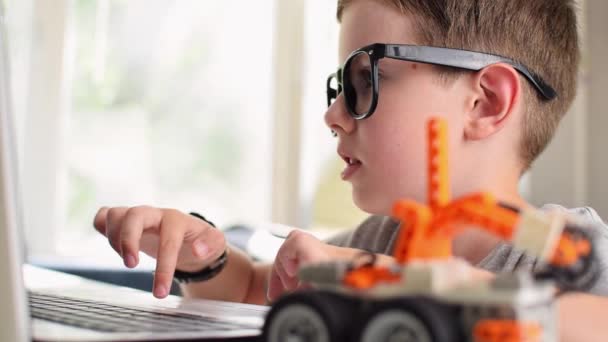 Vue latérale d'un garçon vif à lunettes programme une voiture robot et dactylographier à un ordinateur portable à la maison. Apprendre le codage et la programmation. Robotique, Sciences, Mathématiques, Génie, Technologie, Éducation aux STIM — Video