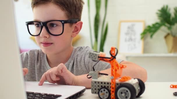 Garoto inteligente sorridente em óculos programa um carro robô usando um laptop em casa. A criança está aprendendo programação e codificação. Robótica, Ciência, Matemática, Engenharia, Tecnologia. Educação STEM — Vídeo de Stock