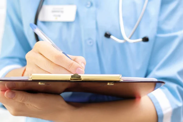 Cropped view of female doctor in uniform with stethoscope holding clipboard and writing records and diagnosis in medical card or patient history in hospital, medical consultation. Close-up.