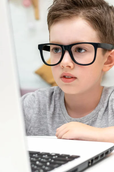 A smart caucasian boy in glasses looks at the laptop screen and learning programming. The child listens to an online lesson at home. Kid play or watch video on computer. Vertical view