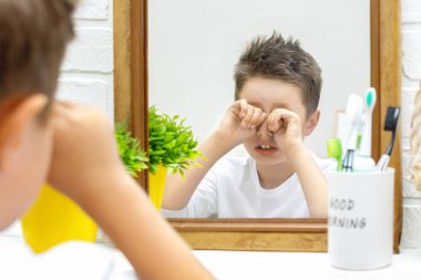 Pijamalı uykulu çocuk aynanın önünde duruyor ve sabahın erken saatlerinde elleri banyoda gözlerini ovalıyor. Çocuk uyandı ve uyumak istiyor. Kötü ruh hali, sabah rutini..