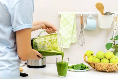 Genç bir kadının sabah beyaz bir mutfakta elmalı ve kivili ev yapımı yeşil ıspanak kokteylini bardağa dolduruşunun arka görüntüsü. Zayıflık için sağlıklı yeme ve diyet kavramı.