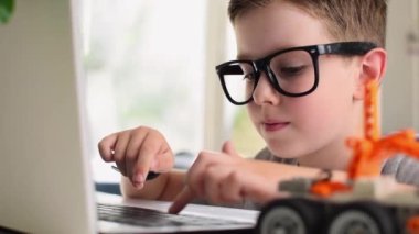 Side view of smart boy in eyeglasses programs a robot car at a laptop at home. Kid learning coding and programming. Robotics, Science, Mathematics, Engineering, Technology, Education. Moving image