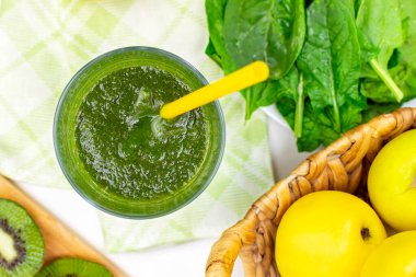Taze ıspanak yapraklı yeşil smoothie, cam içinde elma ve kivi, mutfak masasında sarı pipet, organik sebze ve meyve karışımı, üst manzara. Sağlıklı yaşam tarzı, diyet yeme konsepti.