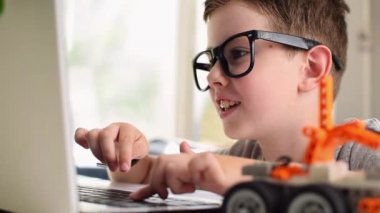 Gözlüklü gülümseyen çocuğun yan görüntüsü evde dizüstü bilgisayarda bir robot araba programlıyor. Çocuk kodlamayı ve programlamayı öğreniyor. Robotik, Bilim, Matematik, Mühendislik, Teknoloji, Eğitim. Resim taşınıyor