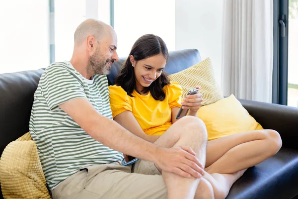 快乐的父亲和年幼的女儿坐在沙发上 一边看智能手机里的东西 一边看电视 — 图库照片
