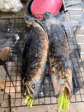 Two whole fish, seasoned with salt and stuffed with fresh green herbs, grilling over hot charcoal on a rustic BBQ rack. Smoke rises from the charred skin, capturing authentic outdoor cooking.