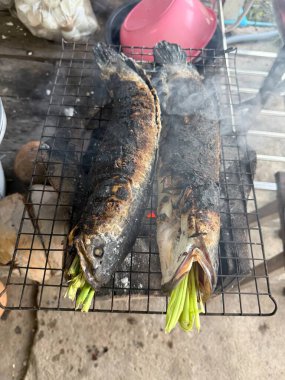 Two whole fish, likely snakeheads, are being grilled over a smoky charcoal stove, stuffed with fresh green herbs, embodying traditional outdoor cooking.