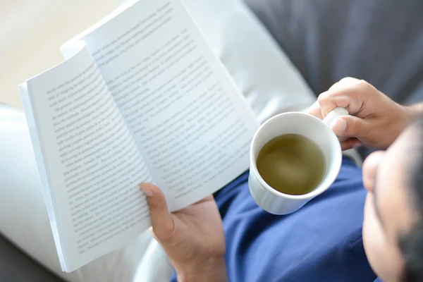 다른 손에 뜨거운 차 컵 책 읽는 남자 — 스톡 사진